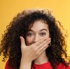 Close-up shot of excited and happy attractive female with curly hair giggling, chuckling and covering mouth as smiling standing amused and joyful as mocking friend over yellow background. Emotions and facial expressions concept